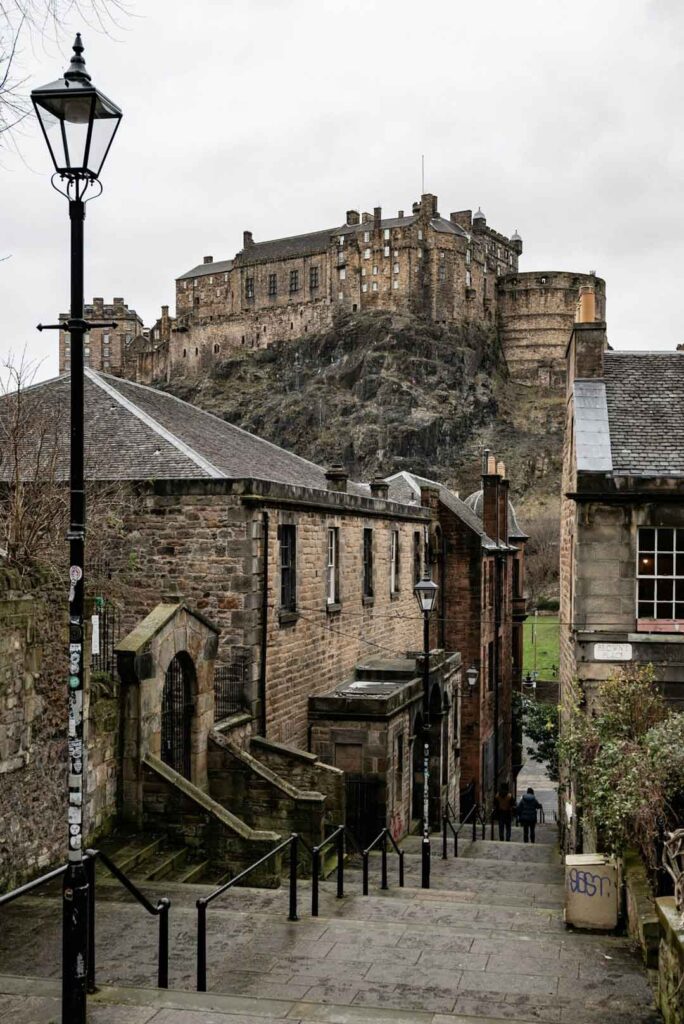 View of Edinburgh Castle perched on volcanic rock, seen from a narrow cobbled street lined with old stone buildings and lampposts.